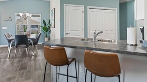 Een moderne keuken met een centraal eiland, barkrukken, een eethoek met stoelen en een groot raam met zonwering.