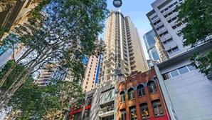 A high-rise building with a 'tbs' sign on top, surrounded by trees and other buildings.