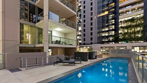 A modern indoor swimming pool with lounge chairs and a glass railing, surrounded by high-rise buildings with balconies and large windows.