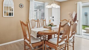 Un coin repas comprenant une table ronde en bois, six chaises en bois, un lustre et une fenêtre avec des stores.
