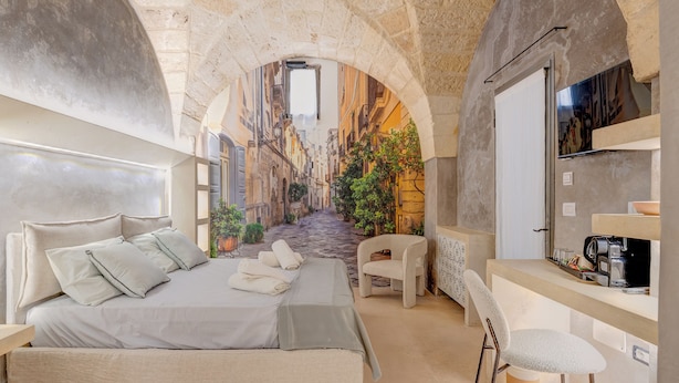A bedroom with a bed, a chair, a desk, and a view of a narrow street through an arched window. - Via Cota 24 (Lecce)