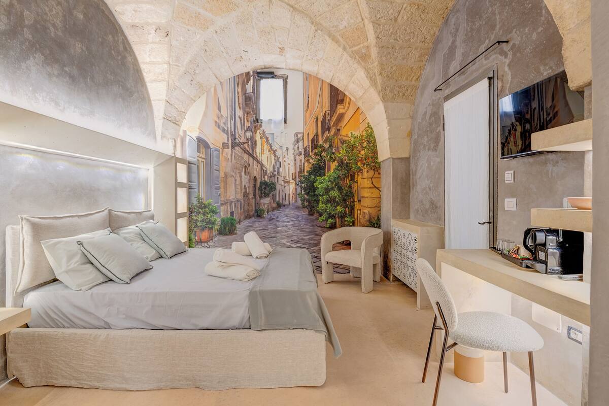 A bedroom with a bed, a chair, a desk, and a view of a narrow street through an arched window.