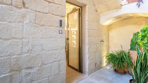 A stone-walled corridor with a door leading to a lit interior, potted plants, and a decorative wall hanging. - Via Cota 24 (Lecce)