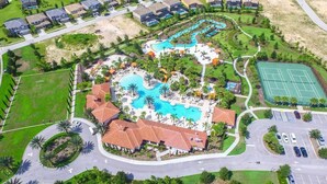 An aerial view of a resort with a central swimming pool, tennis courts, and a parking area.