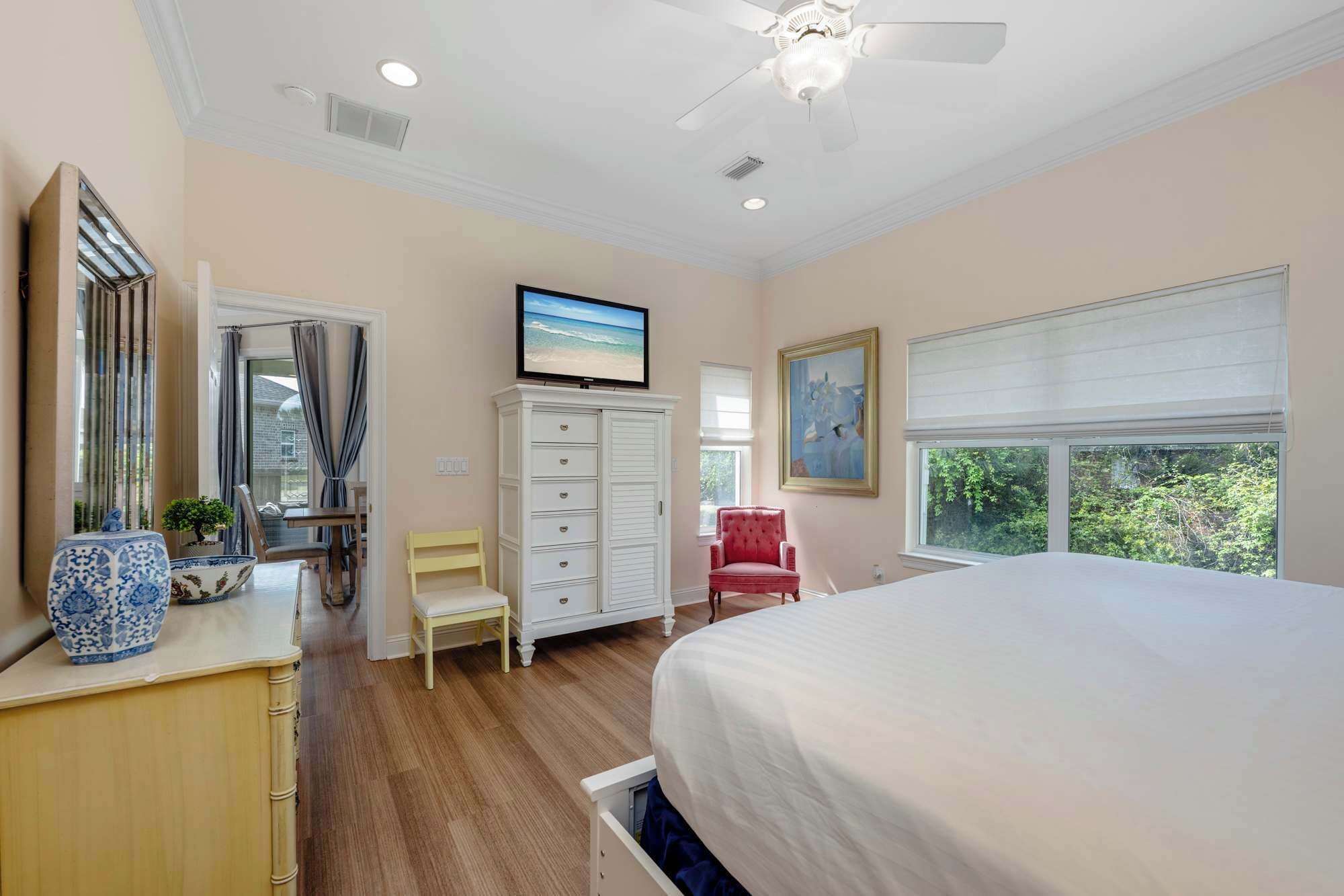 A bedroom with a large bed, a ceiling fan, a TV, a dresser, a chair, and a window with a view of greenery.