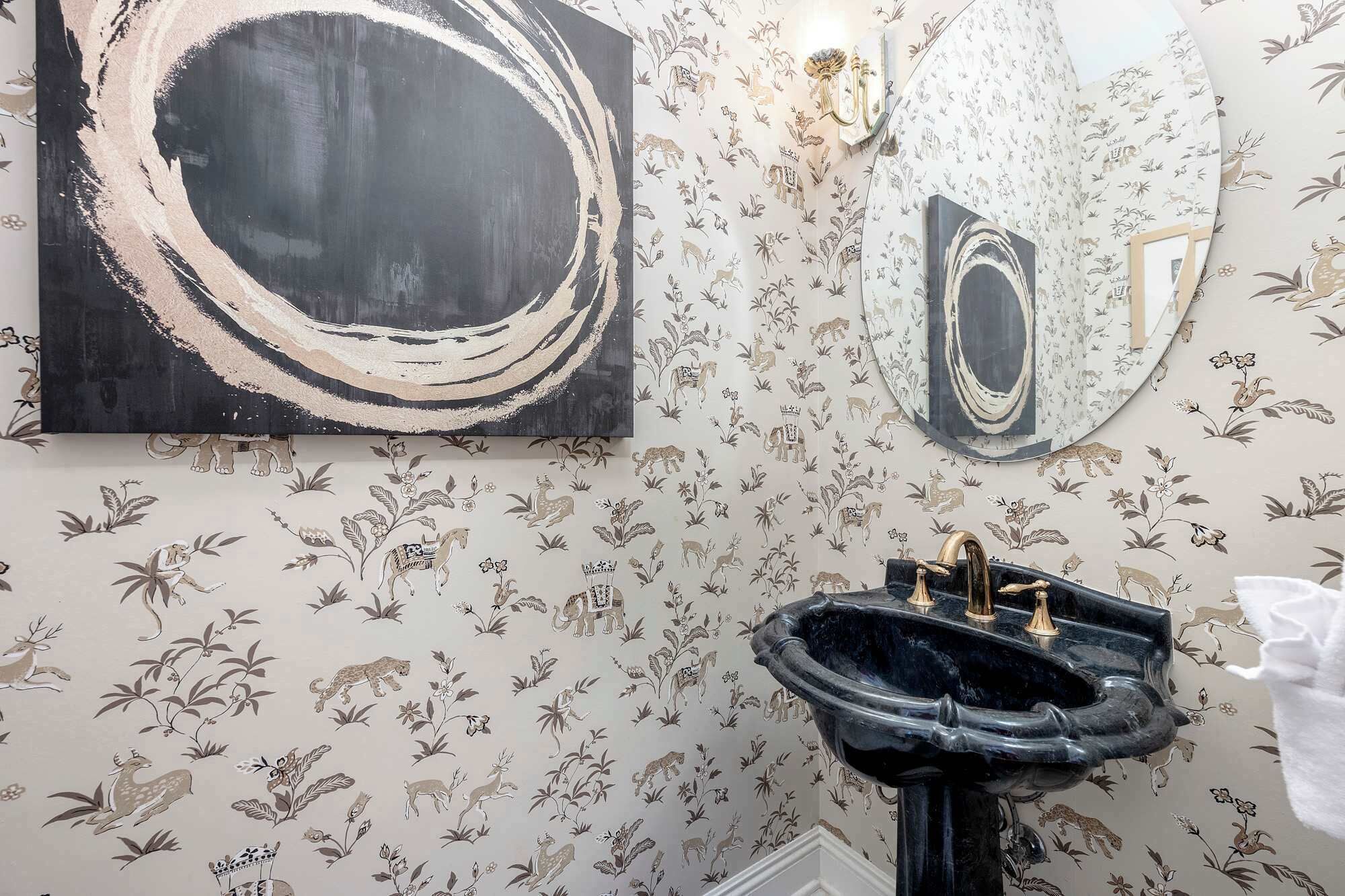 A bathroom with floral wallpaper, a black abstract painting, and a black sink with gold fixtures.