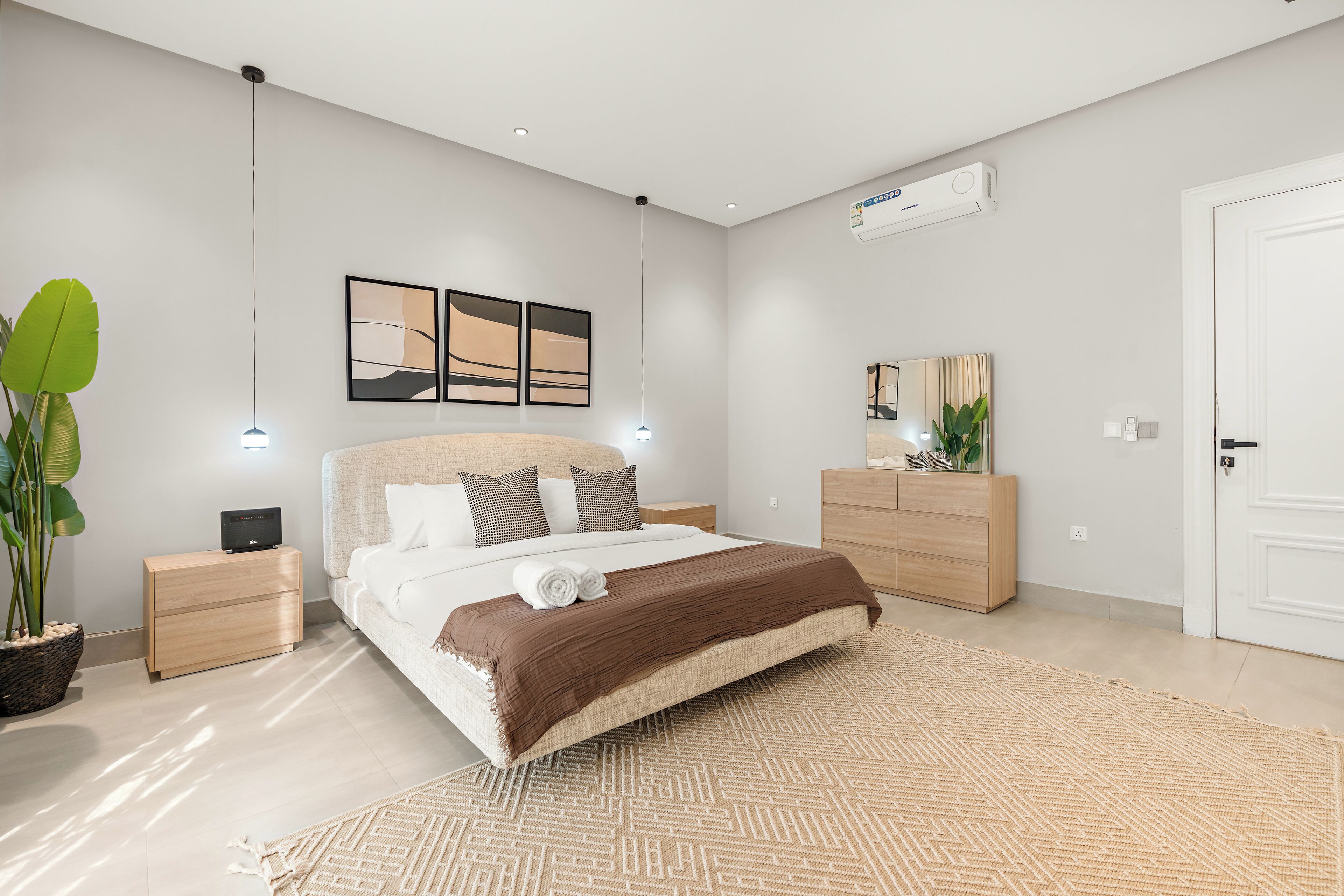 A modern bedroom with a large bed, bedside tables, a dresser, and a potted plant.