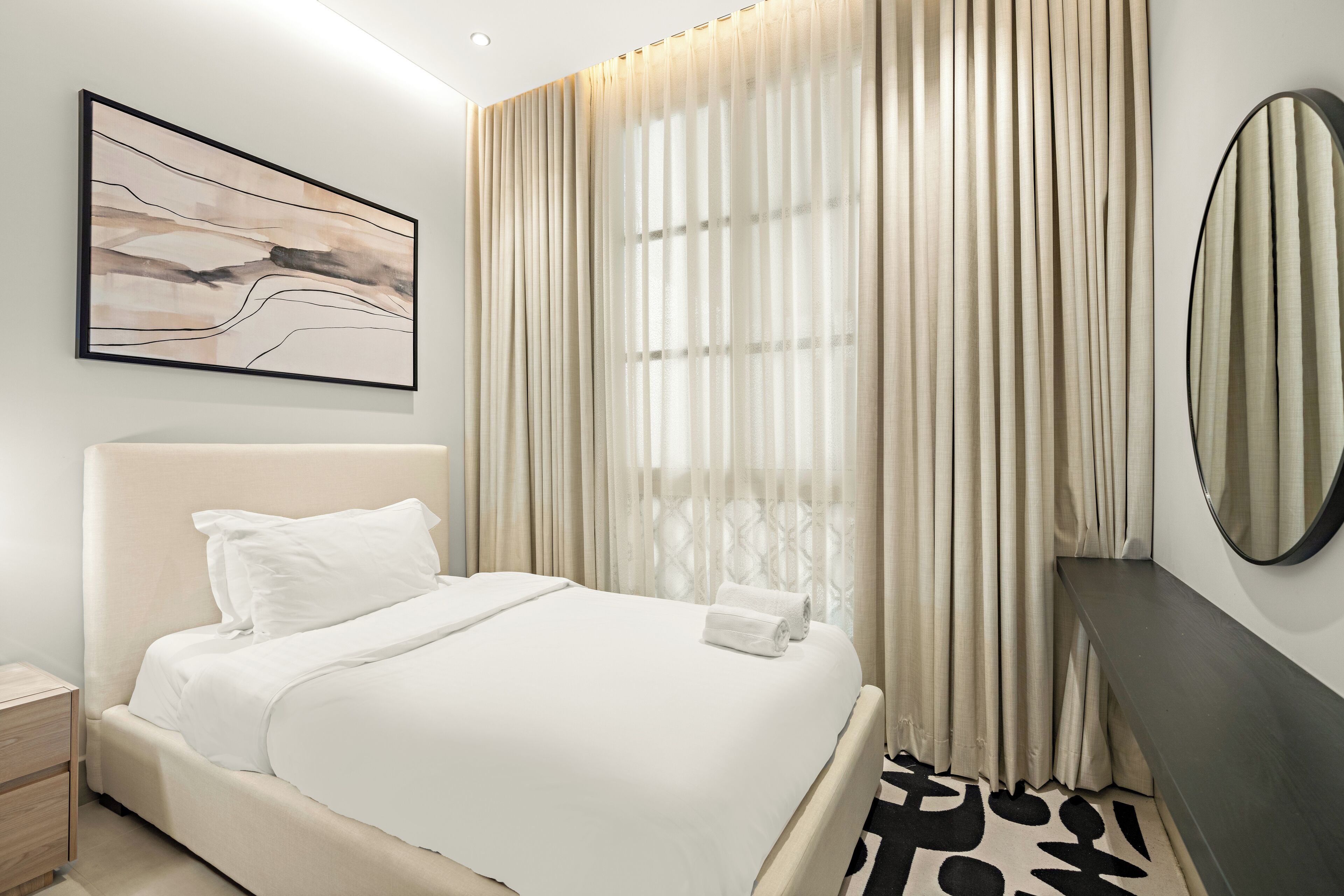 Ein modernes Hotelzimmer mit einem großen Bett, einem Nachttisch, einem wandmontierten Spiegel und einem großen Fenster mit durchsichtigen Vorhängen.