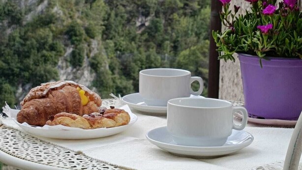 Una mesa con un croissant, dos tazas blancas y una maceta morada, con montañas al fondo.