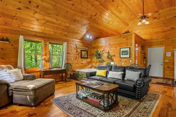 A cozy cabin interior with a wooden ceiling, a fireplace, a sofa, a coffee table, and a window with curtains.
