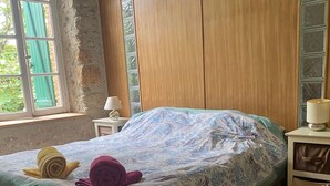 A bedroom with a bed, a nightstand, a lamp, and a window with a view of greenery. - Vacations in the countryside, between the Mediterranean beaches and the Caroux massif, (Murviel-lès-Béziers)