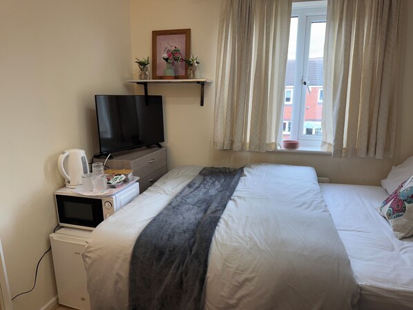 A bedroom with a bed, a television, a small refrigerator, a kettle, and a window with curtains. - Deluxe Double Room in Birmingham (West Midlands)