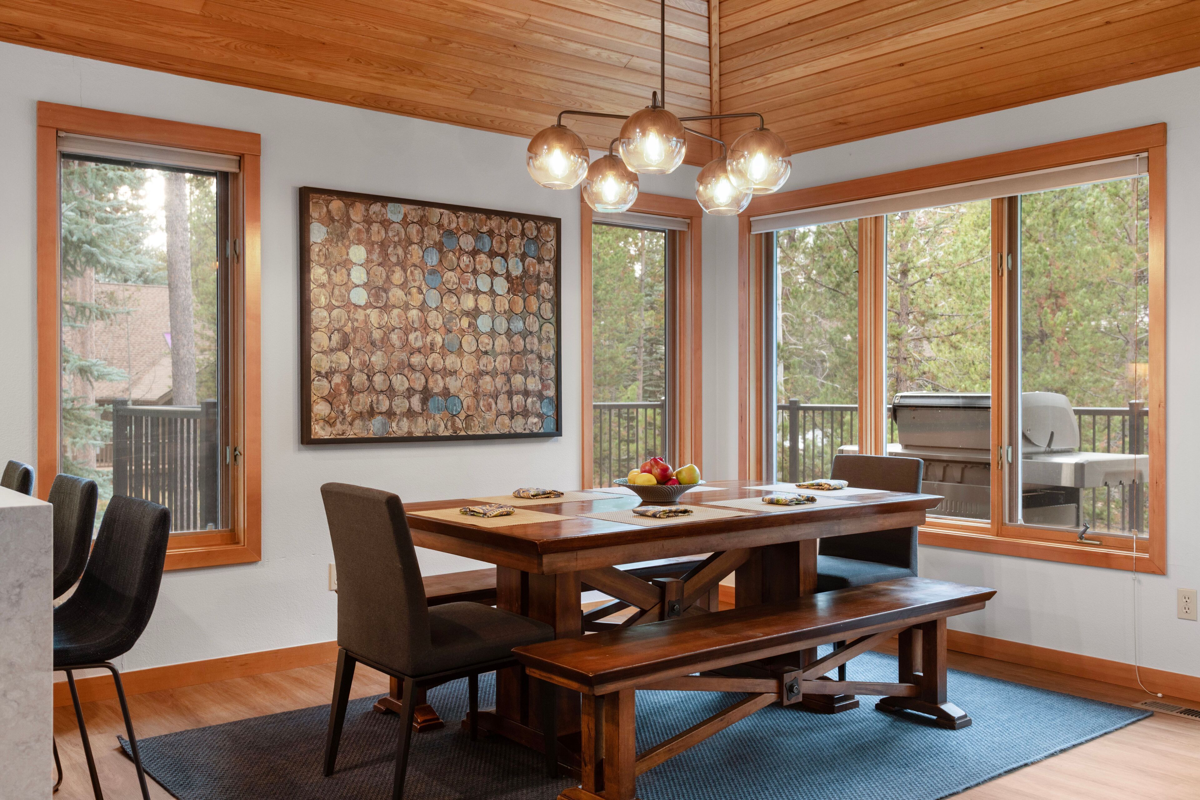 A dining area with a wooden table, chairs, and a bench. There is a large window with a view of trees and a barbecue grill outside.