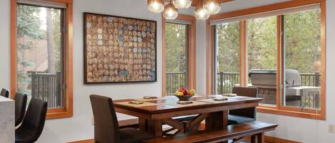 A dining area with a wooden table, chairs, and a bench. There is a large window with a view of trees and a barbecue grill outside.