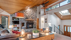 A modern living room with a stone fireplace, a wooden ceiling, and a glass coffee table. - Blue Goose in Sunriver Resort (Sunriver)