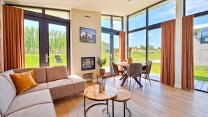 Een ruime woonkamer met een grote bank, een eettafel met stoelen en uitzicht op een groen landschap door grote ramen.
