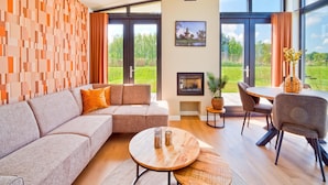 Een moderne woonkamer met een hoekbank, een ronde houten salontafel en een eethoek met een tafel en stoelen. Er is een open haard, een groot raam met uitzicht op groen en een muur met een geometrisch patroon.