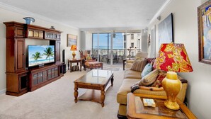 A spacious living room with a large flat-screen TV, a wooden entertainment center, a glass coffee table, and a sofa with decorative cushions. - Gulf and Bay Club- C611 by Tropical Sands Accommodations (Siesta Key)