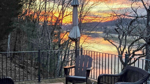 Une terrasse offrant une vue sur un coucher de soleil se reflétant sur une étendue d’eau, le tout entouré d’arbres.