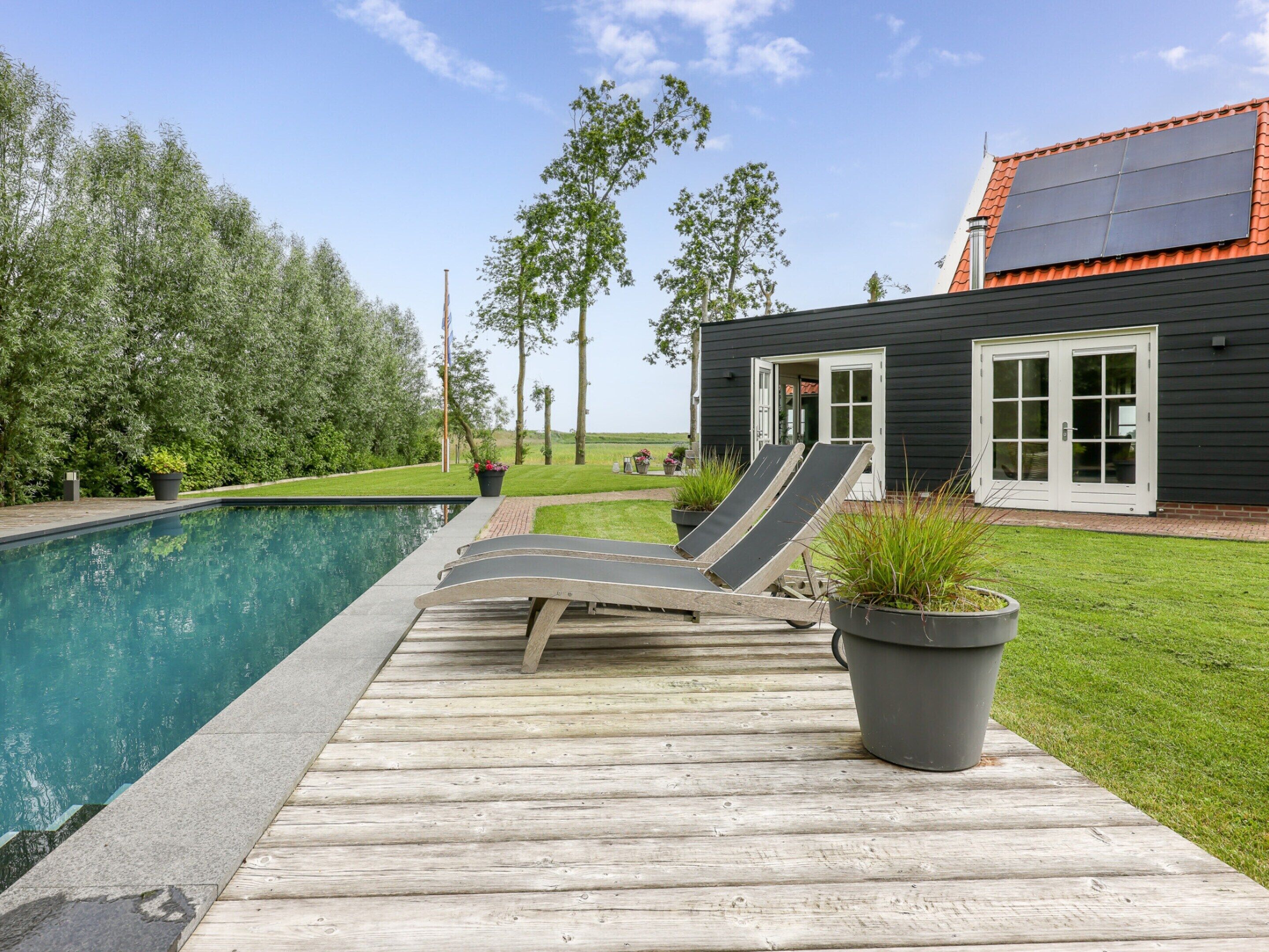 A poolside area with a wooden deck, two lounge chairs, and a house with solar panels and a red roof.