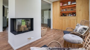 A modern living room with a flat-screen TV mounted on the wall, wooden flooring, wicker furniture, and a built-in wooden shelving unit with books and decorative items. - Holiday Home in Zeeland With Private Pool (Wissenkerke)