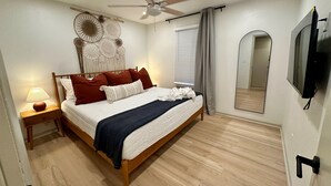 Un dormitorio con una cama, ventilador de techo, lámpara de noche, televisor montado en la pared y un espejo grande.