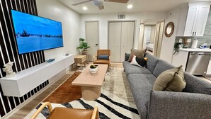 Una sala moderna con un televisor de pantalla plana, un ventilador de techo, un sofá, una mesa de centro y una cocina al fondo.