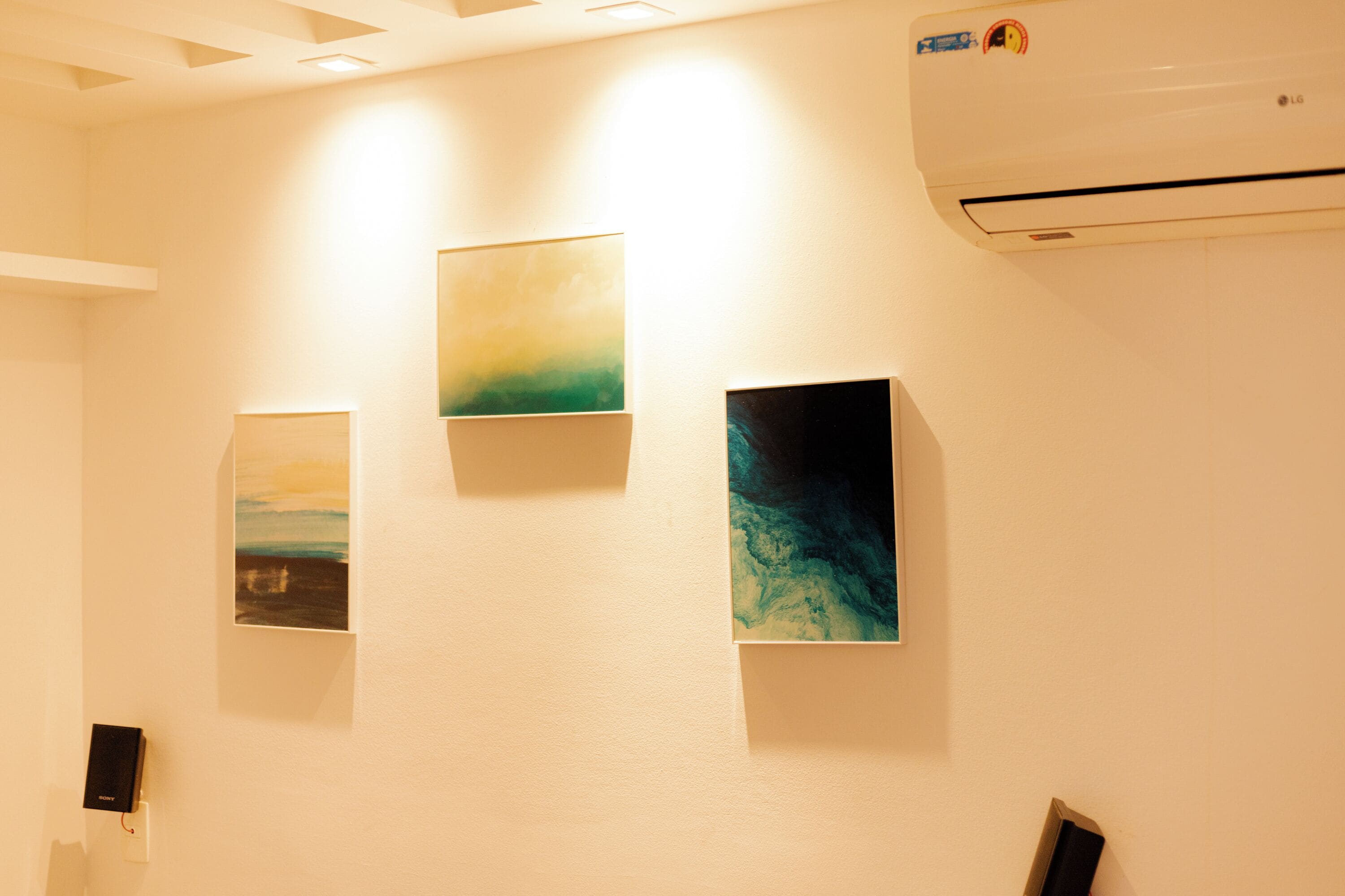 A room with three framed artworks on the wall, a white ceiling with recessed lighting, and an air conditioning unit.