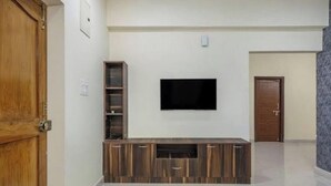 A living room with a flat-screen TV mounted on the wall, wooden entertainment unit, and a door on the left. - Chic 2-bedroom apartment with AC, WiFi in fabulous Hyderabad (Hyderabad)