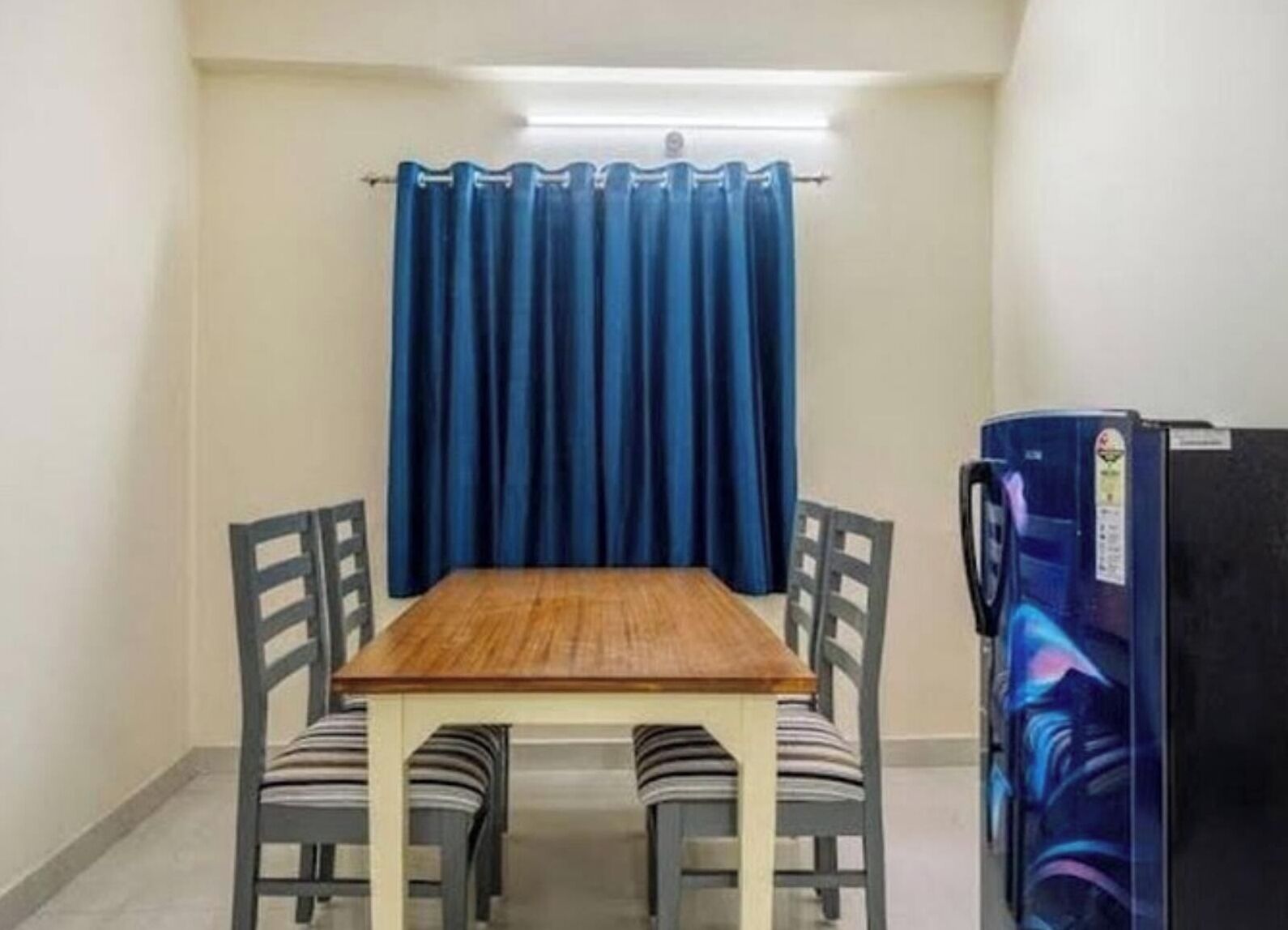 A small room with a wooden table and four chairs, a blue curtain, and a refrigerator.