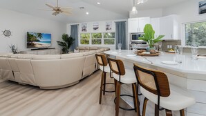 A modern kitchen with white cabinets, a central island with bar stools, a ceiling fan, and a large window with curtains.