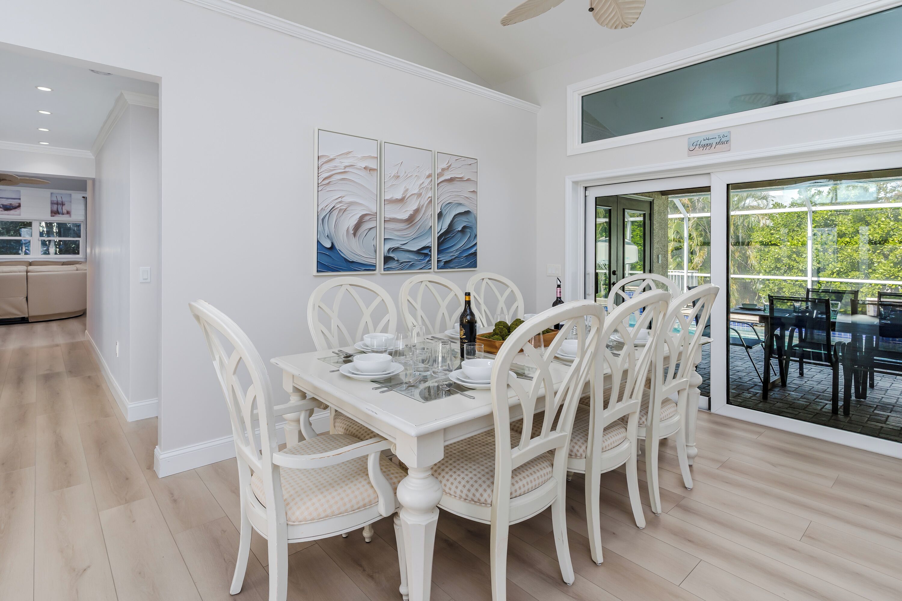 A dining area with a white table set for six, surrounded by white chairs with cushioned seats. There are three wall-mounted abstract paintings above the table.