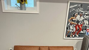 Un canapé en cuir marron, accompagné d’un pouf assorti, dans une pièce dotée d’une fenêtre, d’un tableau et d’une plante en pot.