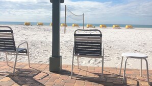 A beachside seating area with metal chairs and a table, overlooking a sandy beach with a volleyball net.