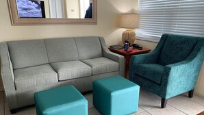 A living room with a grey sofa, two teal ottomans, a teal armchair, a small wooden side table with a lamp, and a framed picture on the wall.