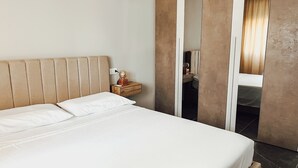 Een net opgemaakt bed met witte lakens, een houten nachtkastje met een wereldbol en een boek, en een spiegel die een kamer reflecteert met een stoel en een tafel.