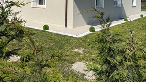Een gebouw met twee verdiepingen, voorzien van een plat dak, witte hekwerk op het balkon en een kleine tuin met struiken en een rots.