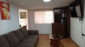 Una sala de estar con sofá, una mesita, televisor y ventana con persianas.