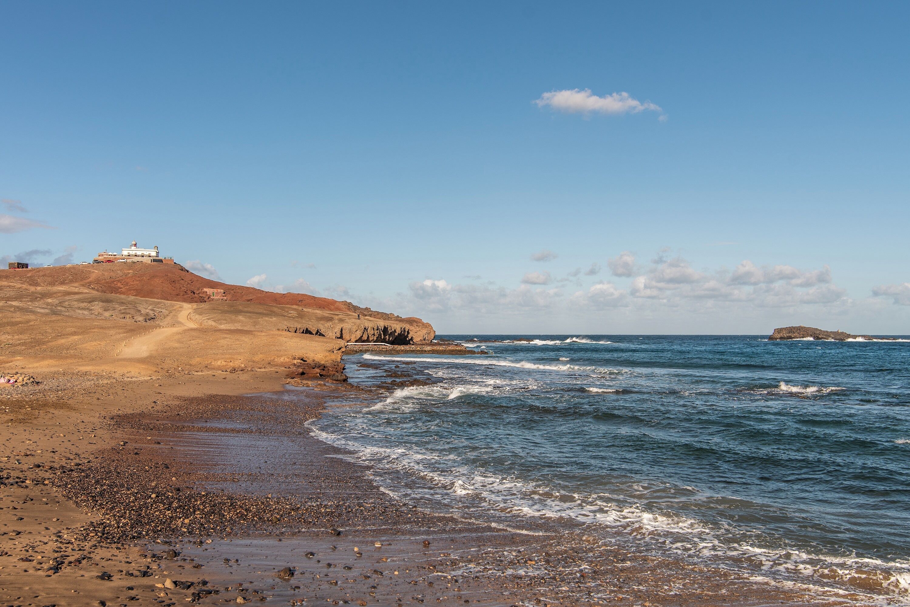 Un paysage côtier avec un phare perché sur une colline, une plage rocheuse et l’océan.
