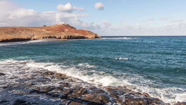 Eine Küstenlandschaft mit felsigem Gelände, einem Leuchtturm auf einem Hügel und dem Meer.