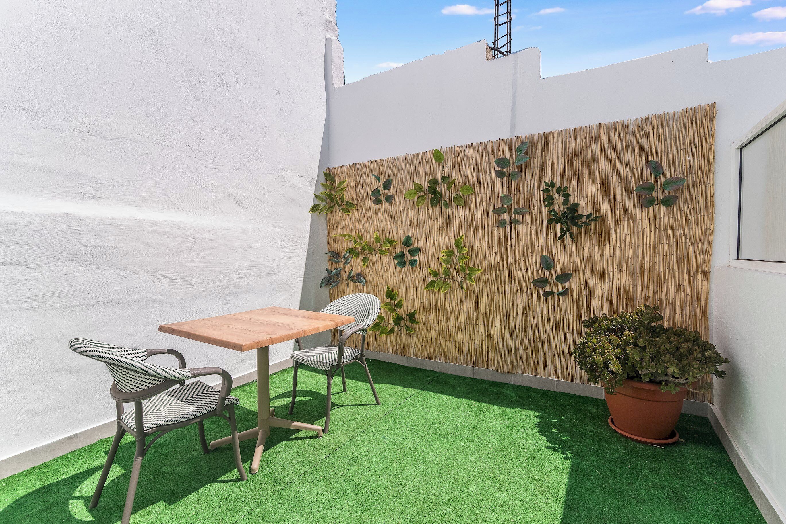 A small patio with a wooden table and two chairs, a potted plant, and a wall decorated with artificial plants.