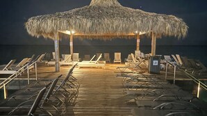 A wooden pier leading to a thatched-roof structure with lounge chairs and a view of the ocean.