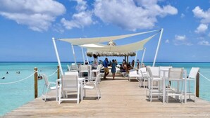 A wooden pier leading to a beachside seating area with white chairs and a thatched-roof structure.