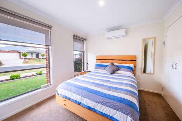 A bedroom with a large window, a bed with striped bedding, a bedside table, and a mirror on the wall.
