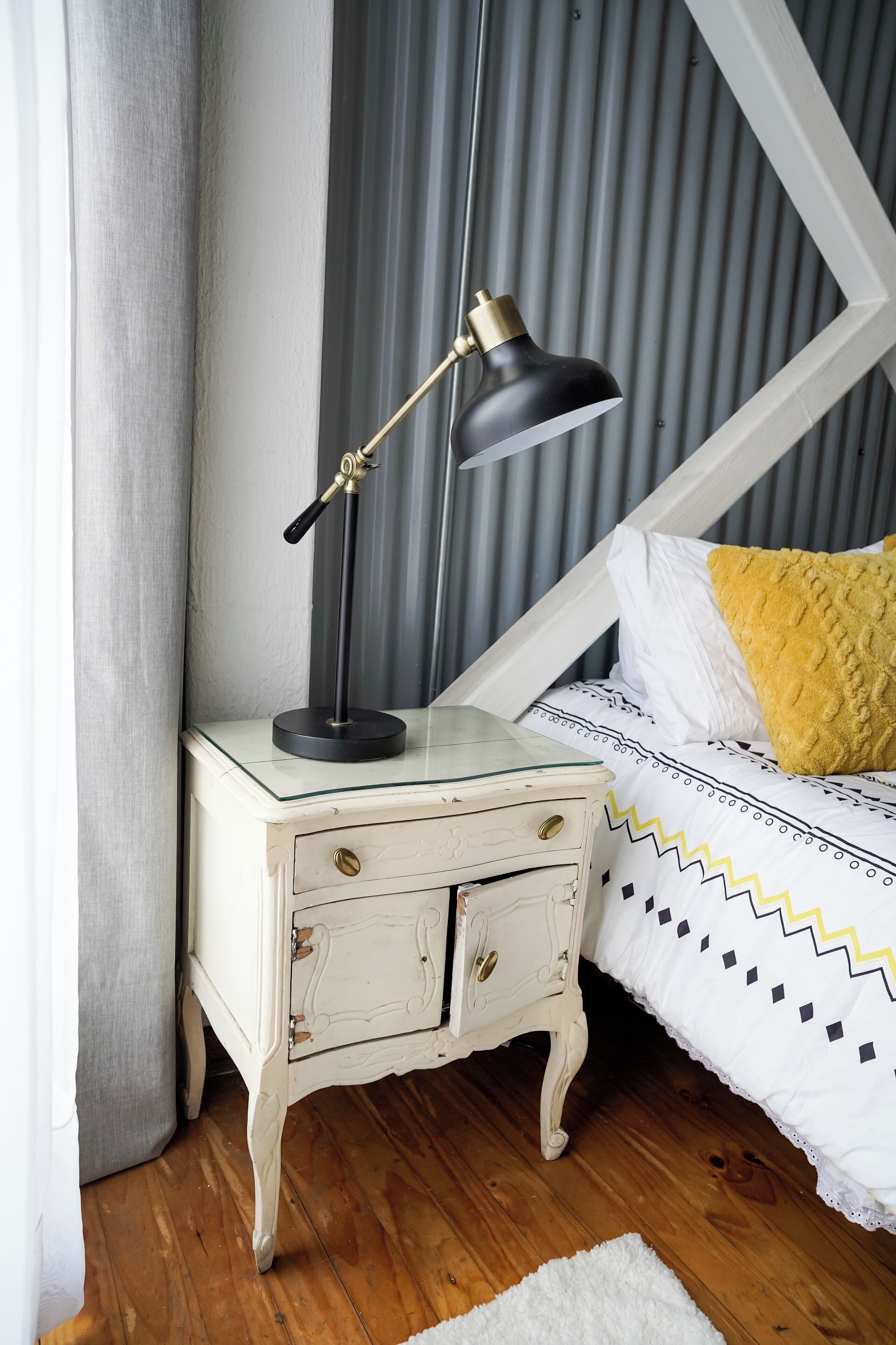 Un dormitorio con una cama blanca, un almohadón amarillo, una lámpara de escritorio negra y una mesita de noche blanca con un cajón.