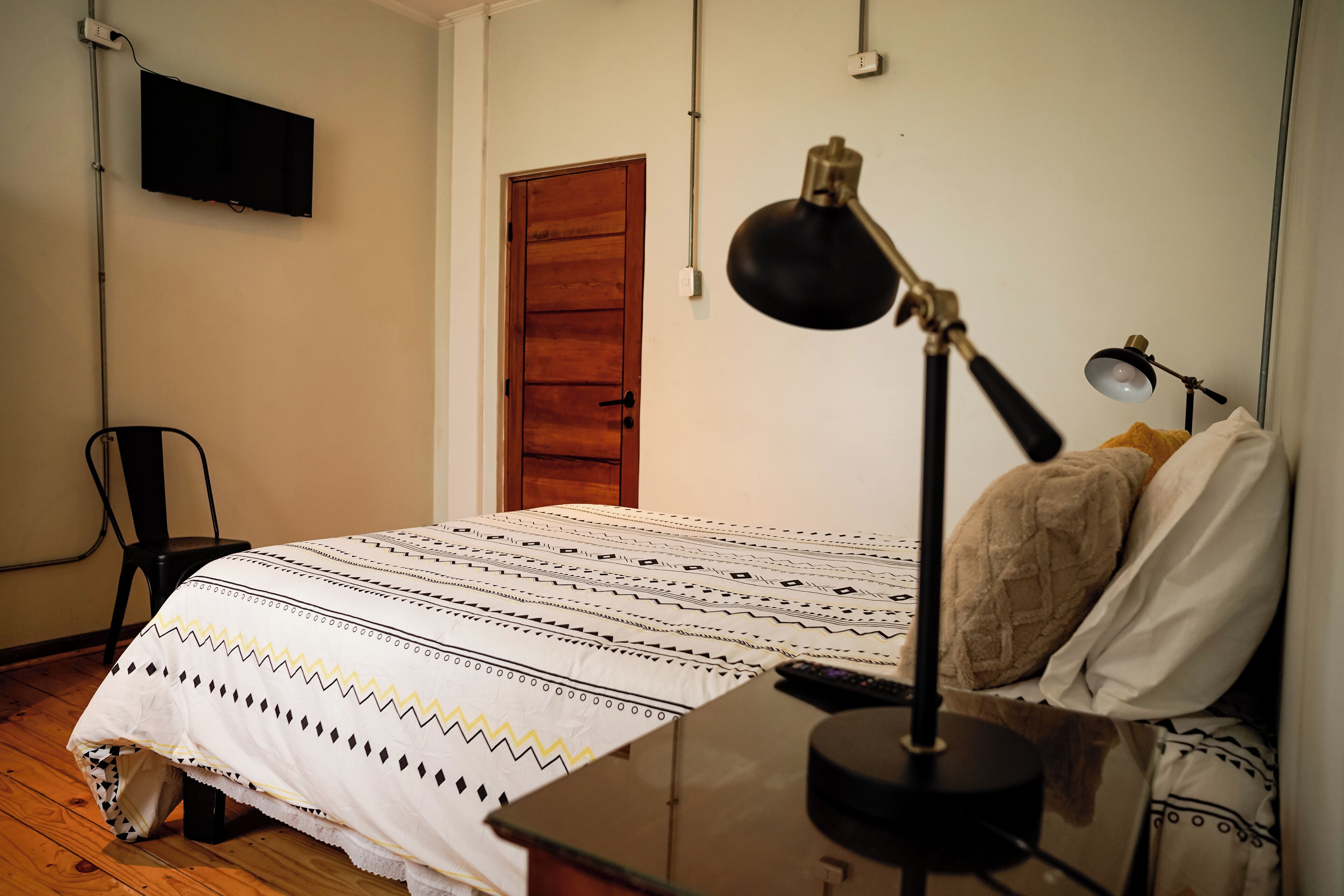 A bedroom with a bed, a desk, a chair, a lamp, and a television on the wall.