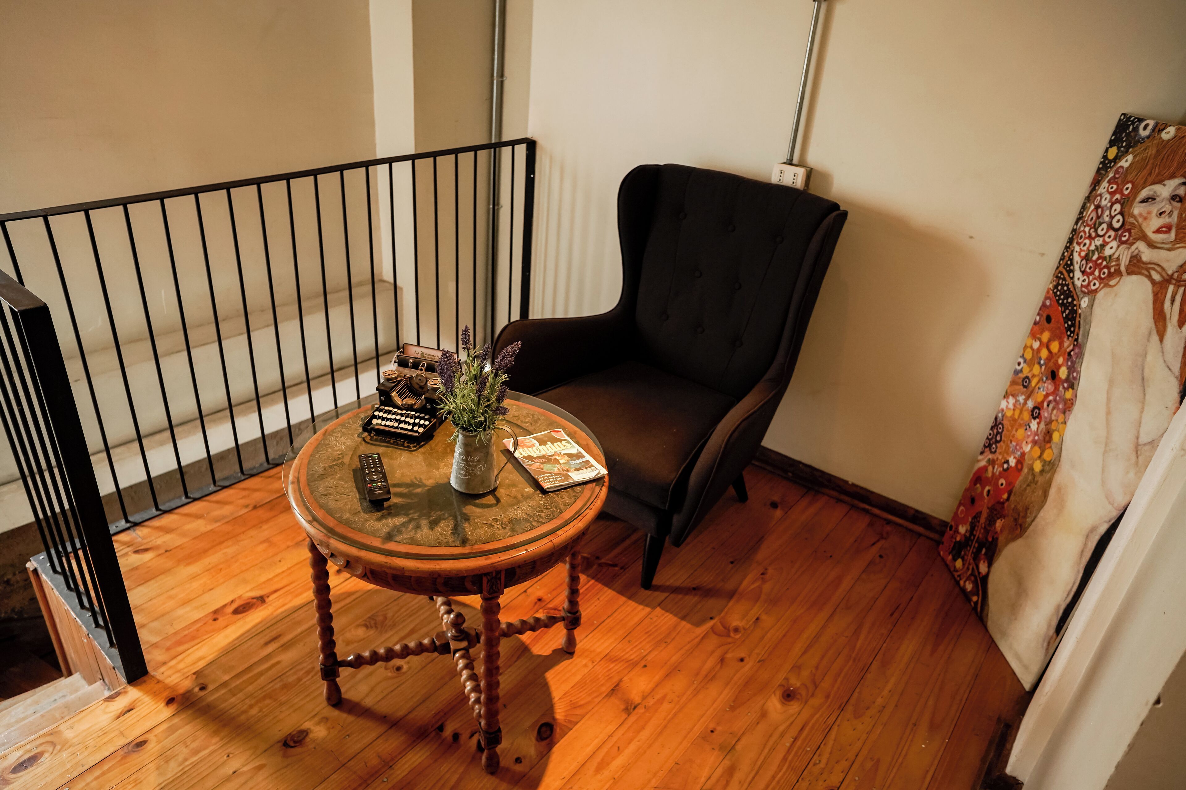 A room with a wooden floor, a round table with a vase and remote controls, a black chair, and a painting on the wall.