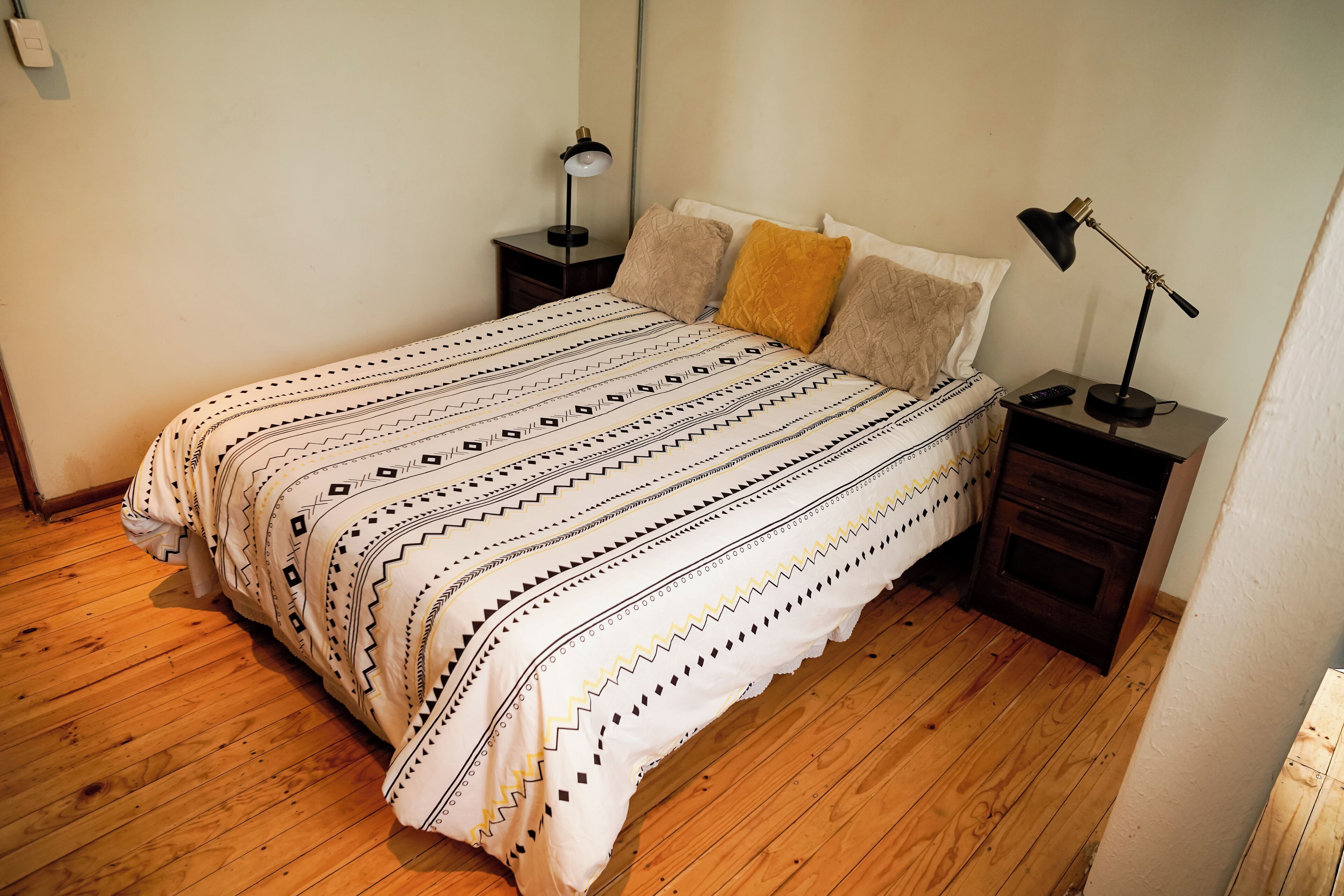 Siististi pedattu sänky kuviollisilla vuodevaatteilla, kaksi yöpöytää, joissa on lamput, ja puulattia.