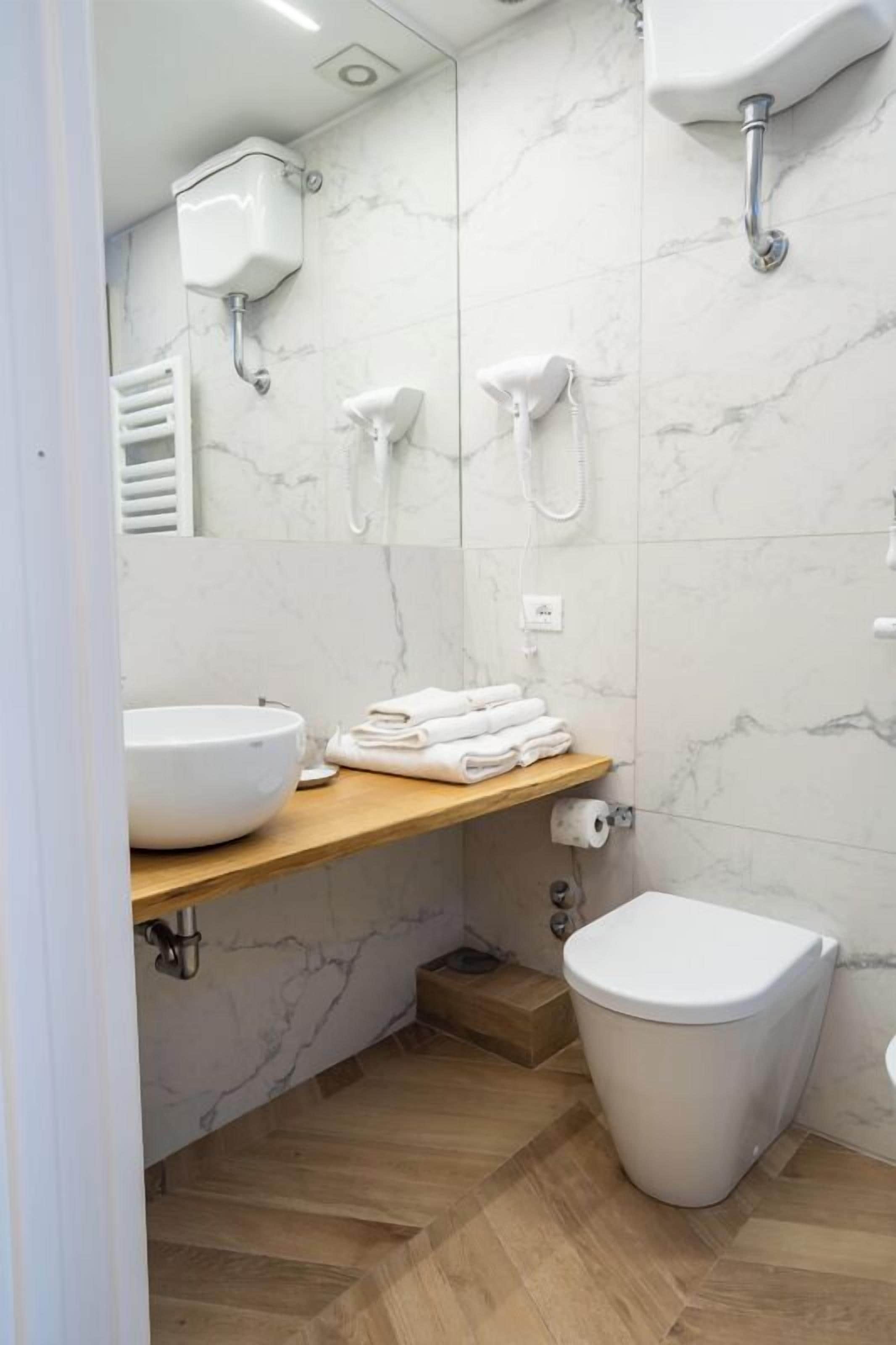 A modern bathroom with a marble backsplash, a wooden vanity, a large white sink, a toilet, and a shower with a handheld showerhead.