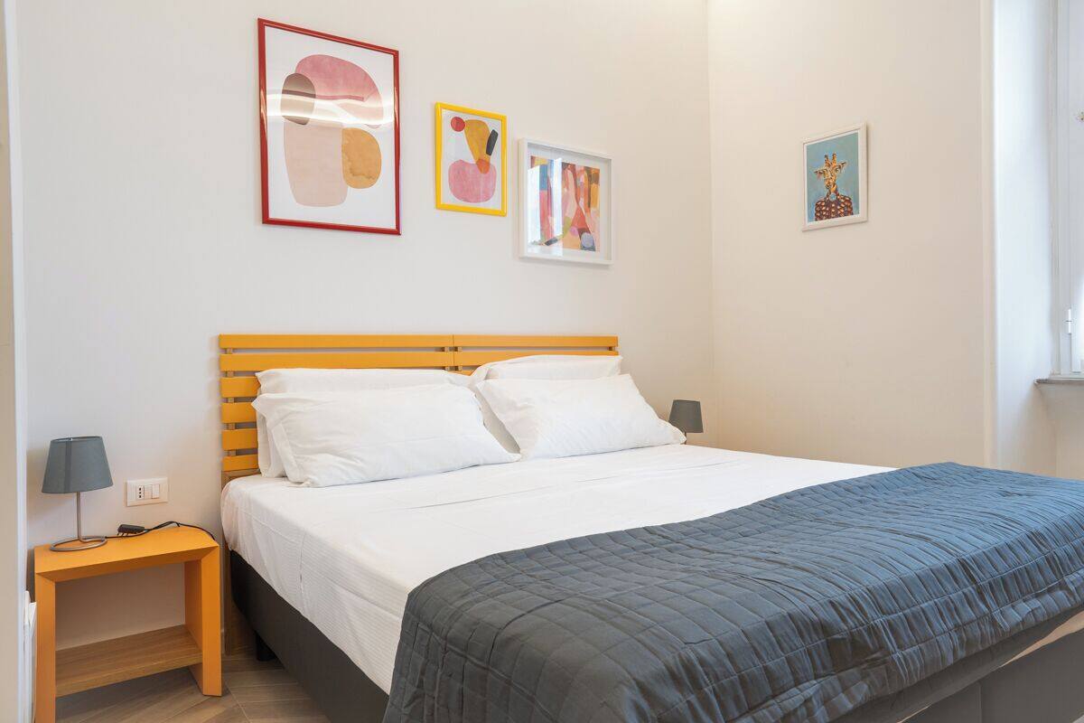 A neatly made bed with a wooden headboard, a bedside table with a lamp, and three framed artworks on the wall.
